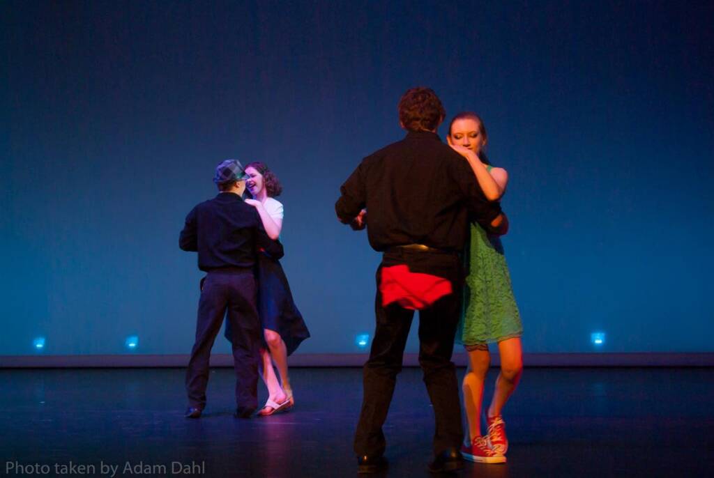 Four people dancing on stage in pairs against a blue background, wearing casual attire. The two pairs are engaged in dance moves, with one couple in the foreground and the other in the background.