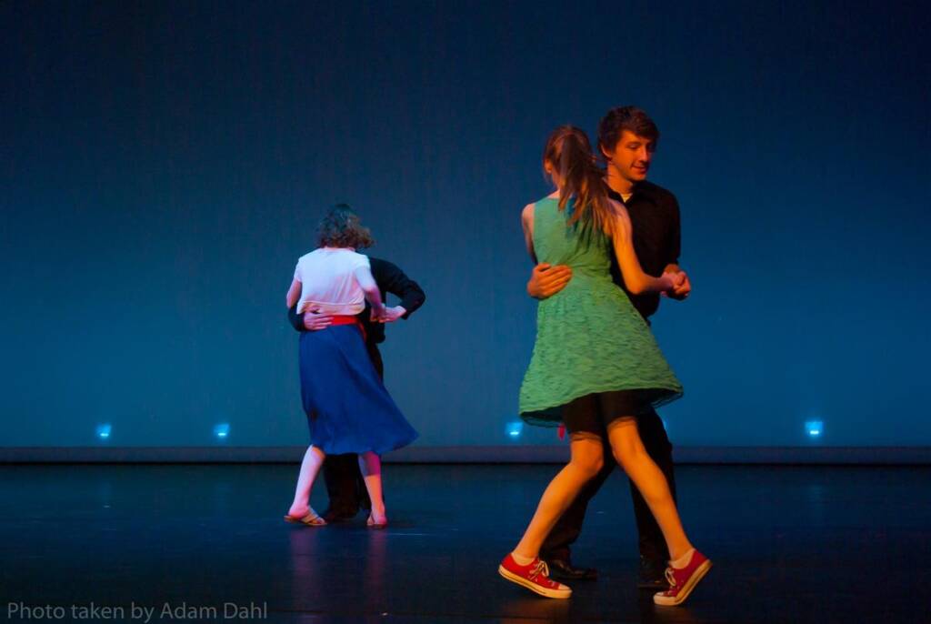 Two pairs of dancers perform on stage. The pair in the foreground includes a woman in a green dress and red shoes dancing with a man in a black outfit. The background pair consists of a woman in a blue dress and a man in dark clothing.