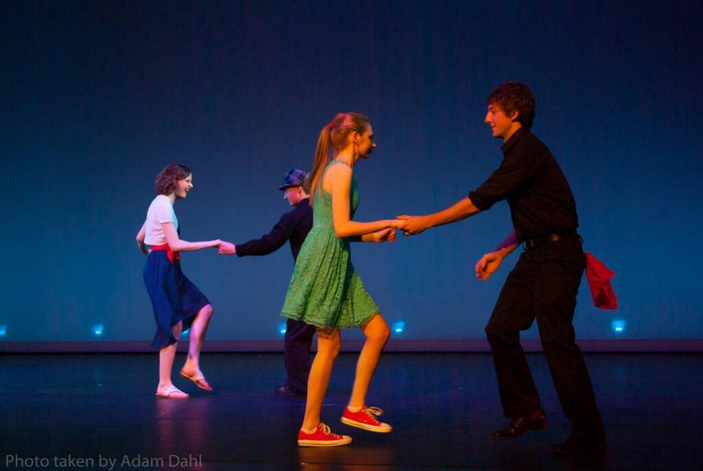 Four people are swing dancing on stage. Two couples hold hands, with the front pair wearing a green dress and black outfit. The rear pair wears a white shirt and black hat. The background is a simple blue, creating a lively dance scene.