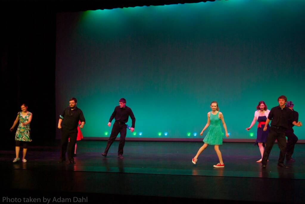 A group of performers on stage dancing in front of a blue-green background. They are wearing colorful attire, including dresses and suits. The lighting casts a vibrant glow, enhancing the energetic atmosphere.