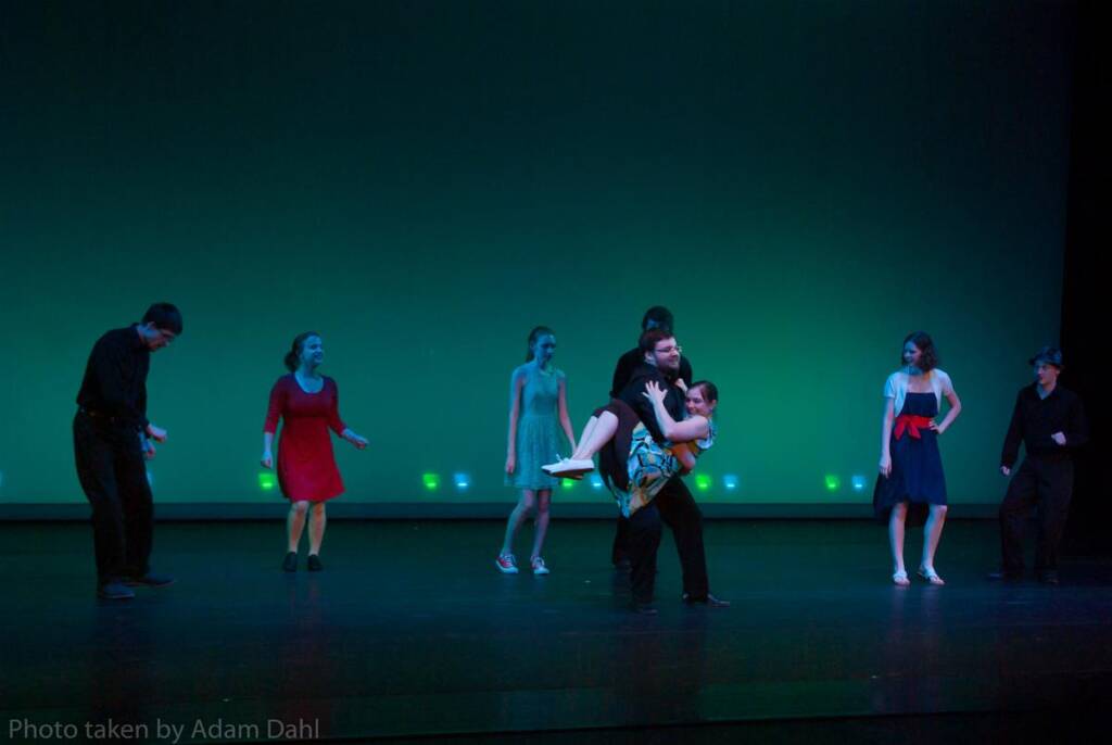 A group of people dance on stage with a blue-green background. One man lifts a woman in a playful pose, while others watch and applaud. They are wearing casual, colorful outfits. The scene is lively and energetic.