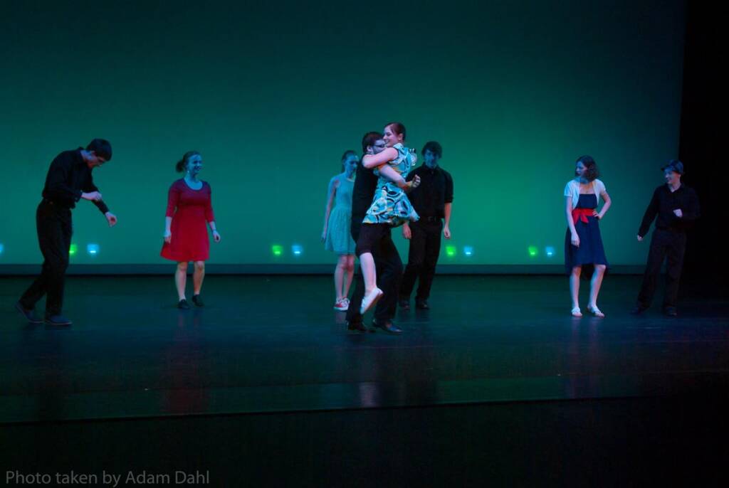 A group of people dancing on a stage with a green-lit background. One person is lifted in the air by another. They're all dressed in semi-formal attire, and the setting appears to be a performance or rehearsal.