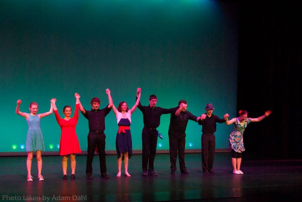 A group of seven people stand on a stage with their hands raised in a bow. They are dressed in various colorful outfits. The background is an illuminated blue-green screen.