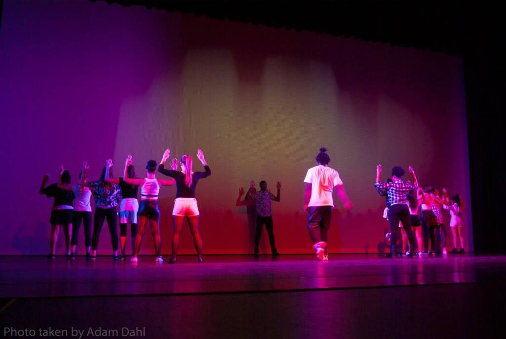 A group of dancers on stage performing in dim, colorful lighting. They are positioned in two clusters, facing a backdrop with their arms raised. The scene has a dynamic, energetic ambiance.