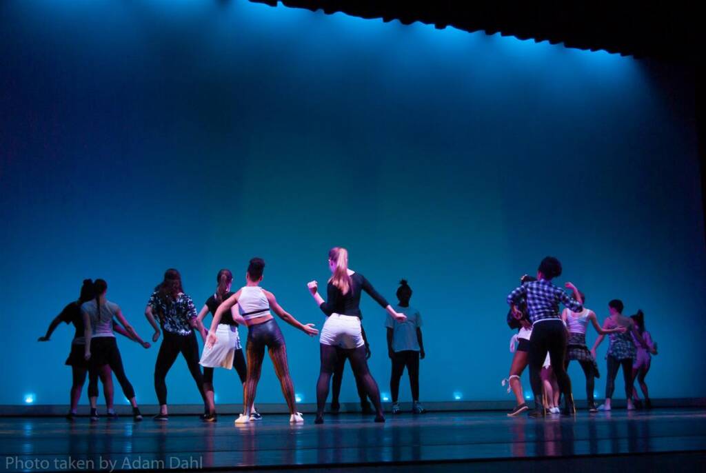 A group of dancers on stage performing against a gradient blue background. They wear casual attire and are in various energetic poses. Stage lights illuminate the scene, creating a vibrant atmosphere.