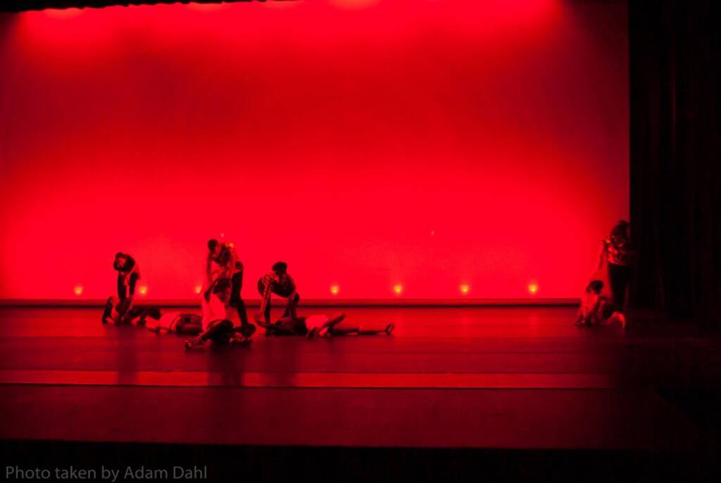 A group of performers on a stage illuminated by a deep red light. Some are standing, while others are seated or lying on the floor. The dramatic lighting casts shadows, creating an intense, theatrical atmosphere.