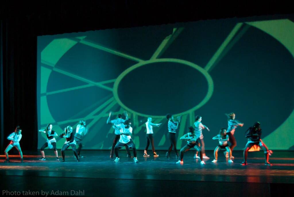 A group of dancers perform energetically on a dimly lit stage. They are arranged in various dynamic poses against a backdrop featuring a large abstract circle design with intersecting lines.