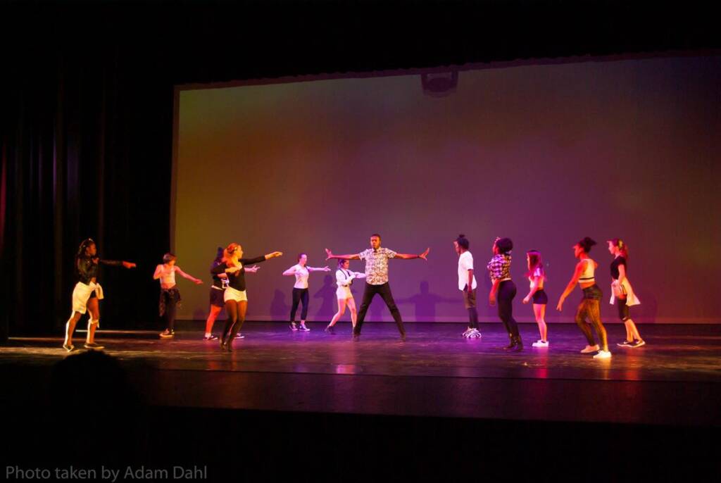 A group of performers on stage, dancing under colorful lights. They are in various poses, some pointing and others extending their arms. The background is a plain, illuminated screen.