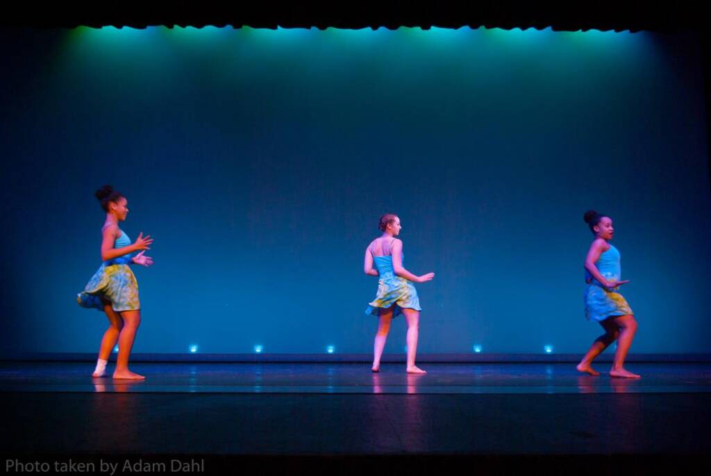 Three dancers perform on stage in colorful outfits against a blue backdrop. They are barefoot, mid-movement, each facing a slightly different direction, creating a dynamic and synchronized composition.