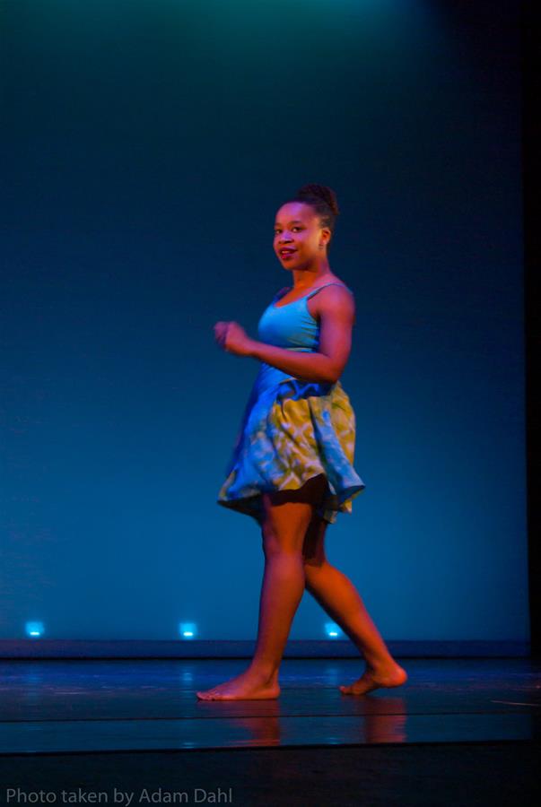 A dancer on stage wearing a blue and green dress, performing under moody blue lighting. She is barefoot and facing slightly to the side, with one arm raised as she moves gracefully.