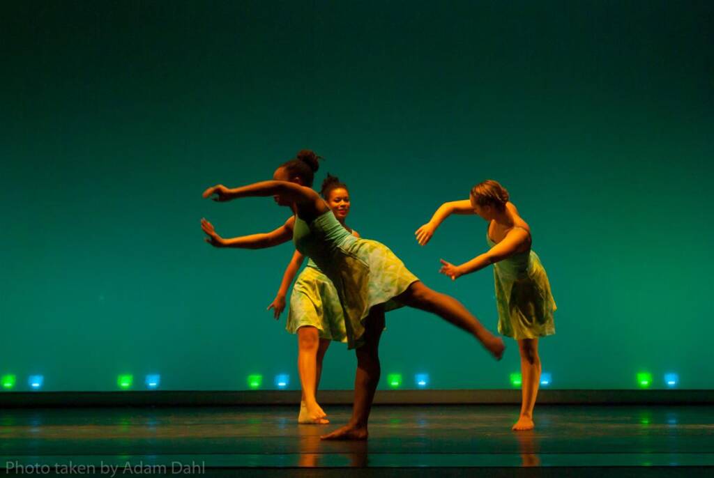 Three dancers on stage perform in synchrony against a vibrant green background. They wear light, flowy dresses, and their movements are dynamic and expressive, capturing a moment of graceful balance.