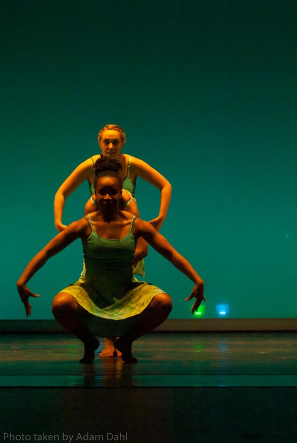 Two dancers on stage perform with arms outstretched and knees bent, wearing green dresses. The background is illuminated with a blue-green hue, creating a dramatic atmosphere.