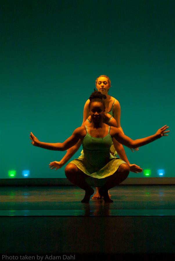 Two dancers pose in coordinated positions on stage. The foreground dancer crouches with arms extended, while the other stands behind, mimicking the pose. They wear matching green dresses. The background is illuminated in shades of green.