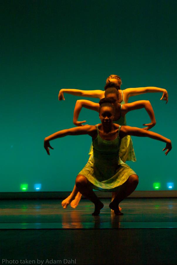 Three dancers in green outfits perform on stage, aligned behind each other. The front dancer is crouched with arms bent and raised, creating a layered visual effect. The background is a vibrant green with subtle colorful lights near the floor.