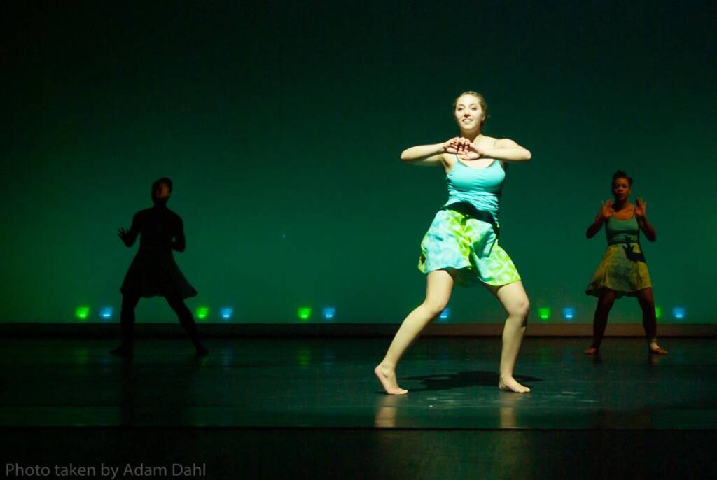 A dancer in a blue and green outfit performs on stage under green lighting, with two dancers in the background. The stage is dimly lit, creating silhouettes of the background dancers.