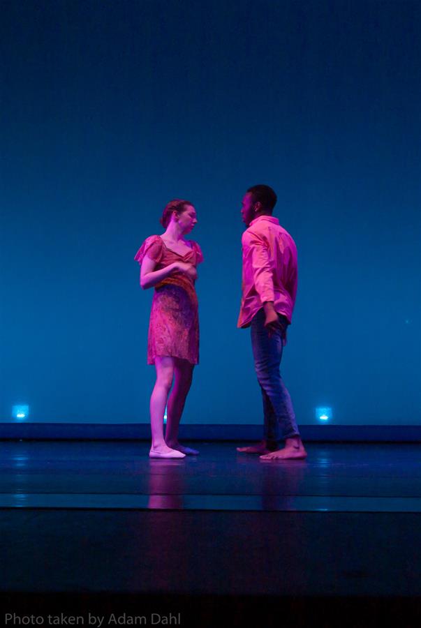 Two dancers are performing on stage against a blue-lit backdrop. They face each other, wearing casual attire; the dancer on the left is in a dress, and the one on the right is in a shirt and jeans. The stage is dimly lit.