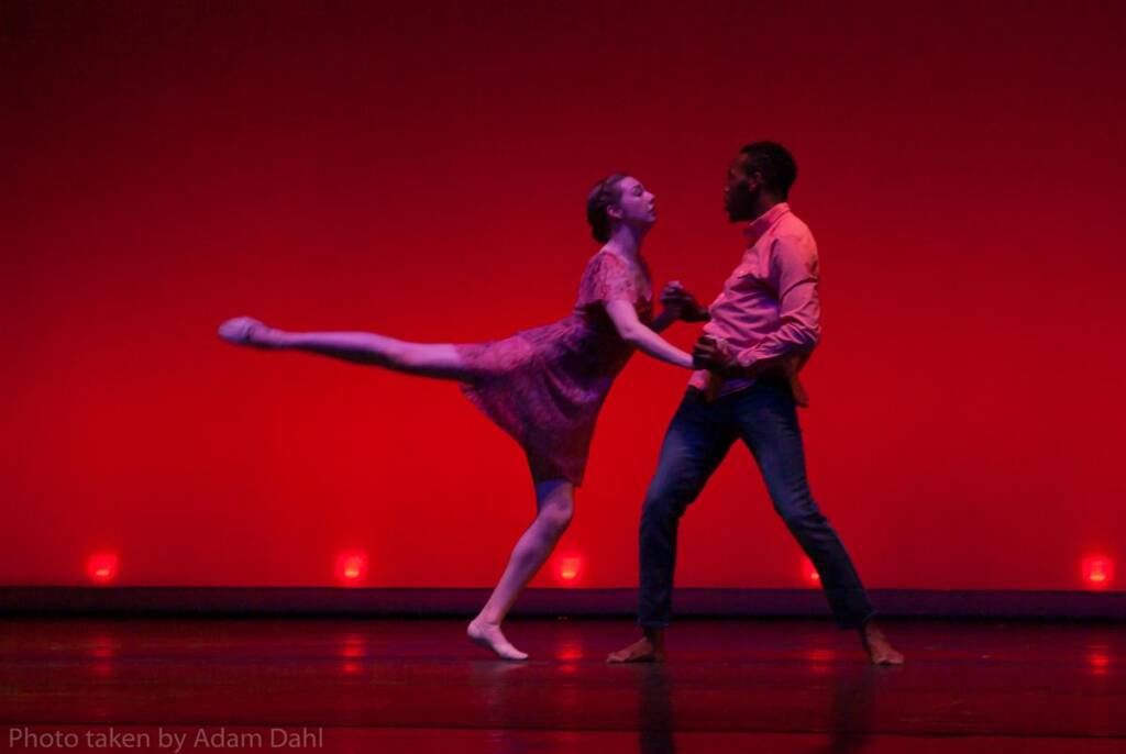 A dancer in a dress extends her leg gracefully while holding hands with a male partner in a shirt and jeans on a stage. The background is illuminated with a deep red hue, adding drama to the performance.