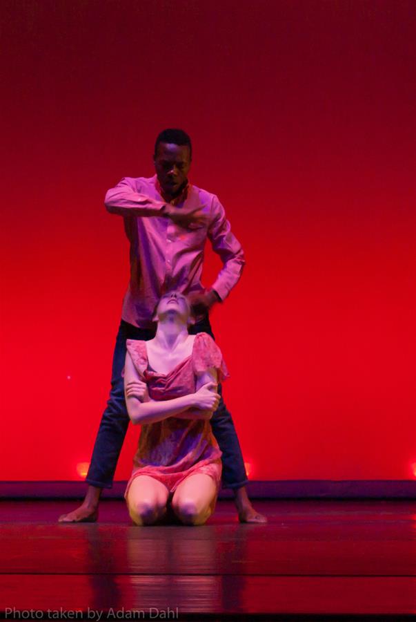 Two dancers perform on stage with a red backdrop. One dancer kneels, wearing a red outfit. The other, standing behind, wears a pink shirt and black pants, with an arm across their chest, conveying an emotional expression.