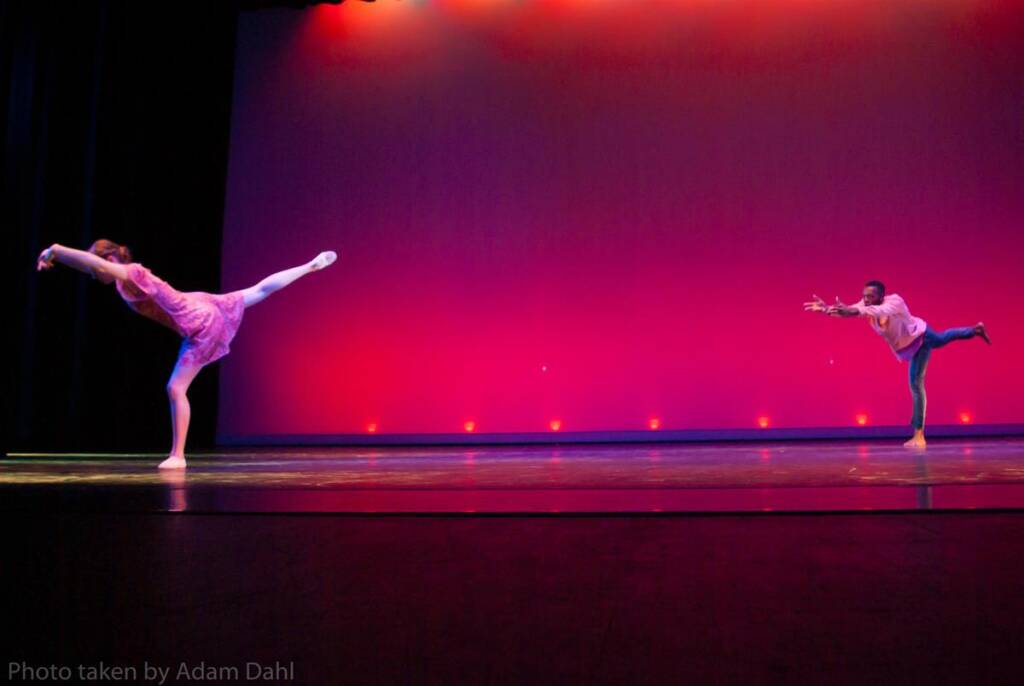 Two dancers perform on stage. One stands on one leg with the other extended back, and the other mirrors the pose from afar. The backdrop is illuminated in red and orange hues, creating a vibrant atmosphere.