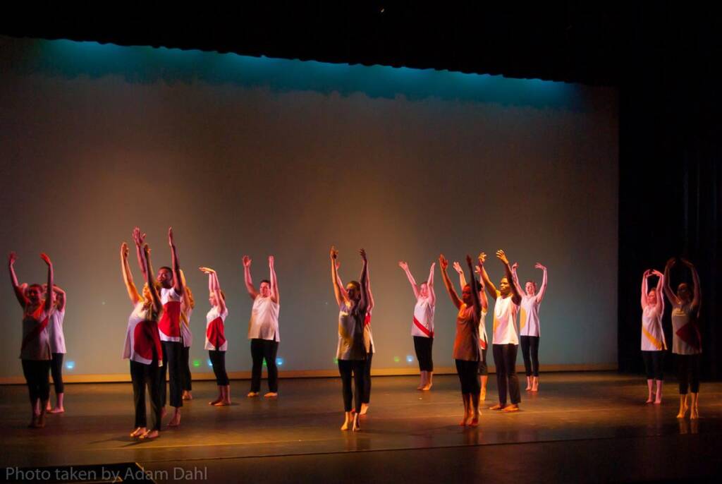 A group of dancers in colorful costumes performing on a dimly lit stage. They are arranged in a formation with their arms raised in the air. The background is a plain, softly lit wall.