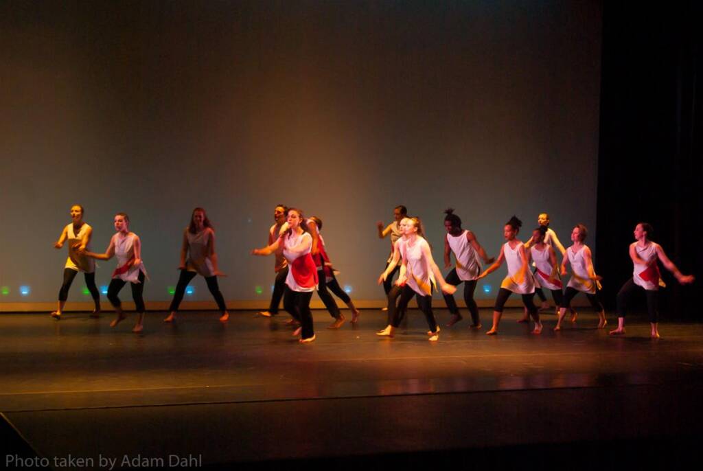 A group of dancers performs energetically on stage, all wearing sleeveless tops with red and white patterns and black pants. The stage is dimly lit, casting shadows behind them, against a plain background.