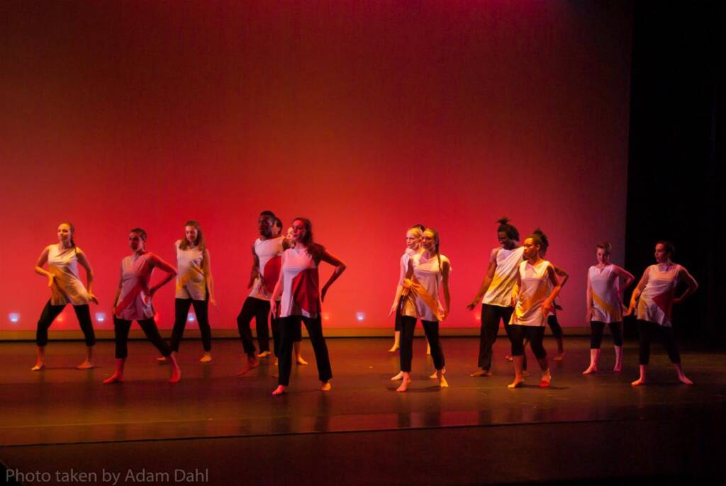 A group of dancers performs on stage, wearing matching white tops and black pants. The stage is lit with a reddish-orange backdrop, creating a warm ambiance. The dancers are in various dynamic poses.