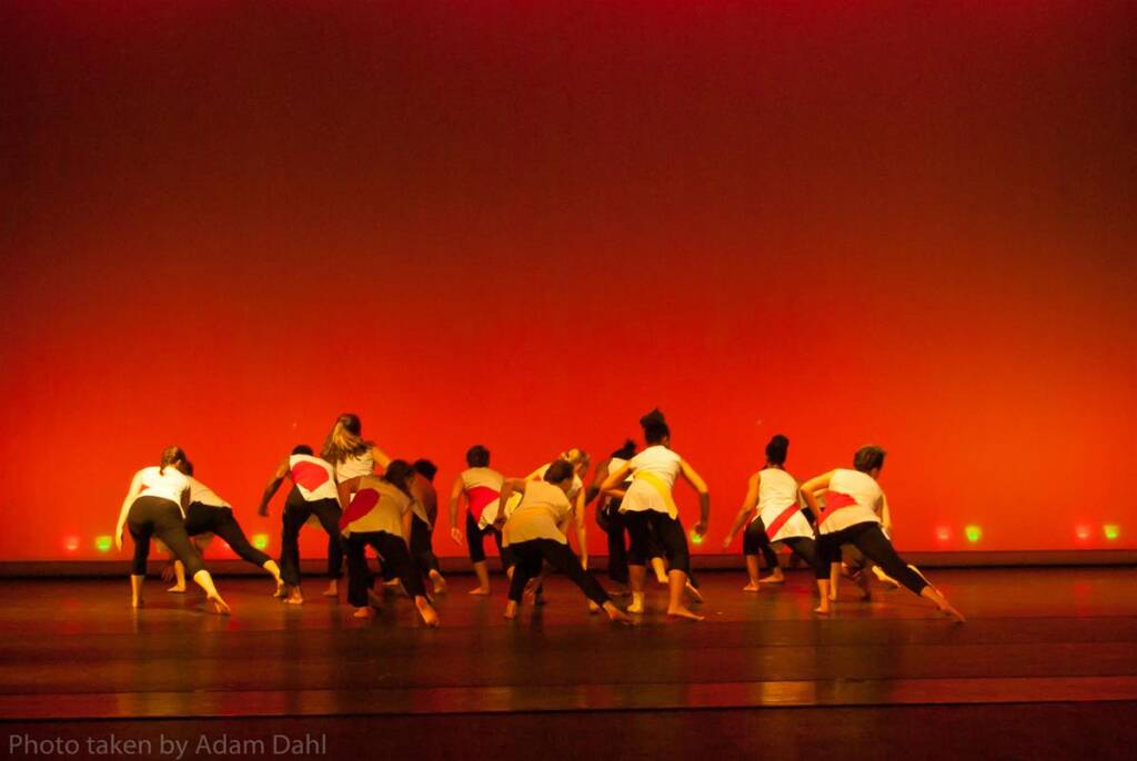 A group of dancers perform on stage, facing away from the audience, under dramatic red lighting. They wear white tops with splashes of color and black pants, moving energetically in a coordinated formation.