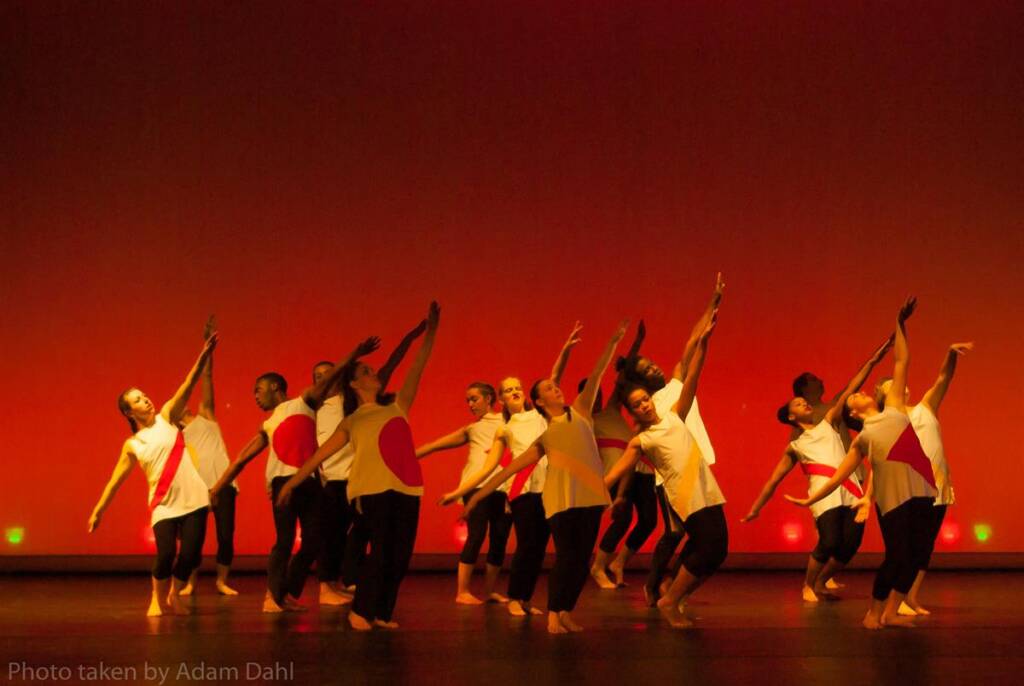 Dancers in white tops and black pants perform a synchronized routine on stage against a red background, with some dancers extending arms upwards. The lighting casts warm tones on the scene.