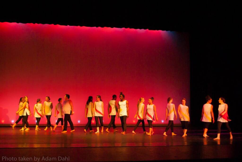 A group of dancers performs on a stage with a gradient red and purple backdrop. They wear white tops and dark pants, moving across the stage in an organized formation. The lighting and colors create a vibrant atmosphere.