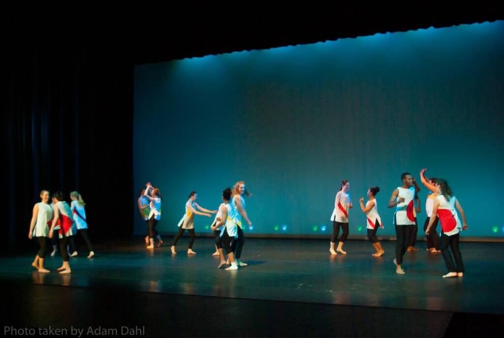 A group of dancers on stage perform a routine. They're wearing outfits with red and white accents. The stage lighting casts a blue glow in the background. The dancers are spaced out, some mid-movement with arms raised.