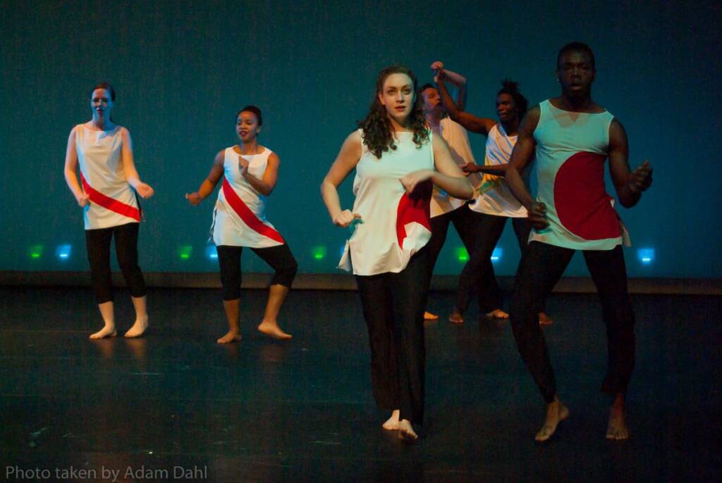 A group of six dancers performs on stage wearing matching white tops with red and blue curved designs. The background is dark, with colorful lights illuminating the performers.