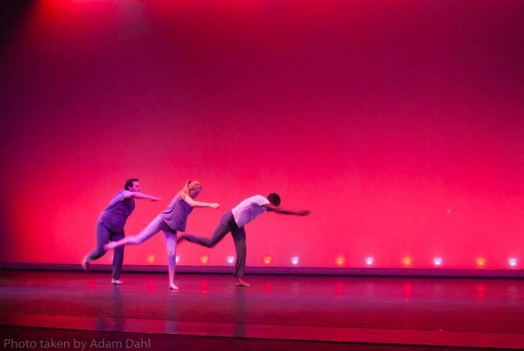 Three dancers perform on stage against a red-lit backdrop. They are in mid-motion, leaning forward with one leg extended behind them. The stage is dimly lit with small lights at the base.