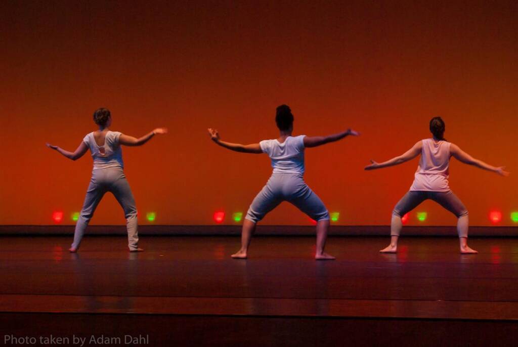 Three dancers in white outfits perform on stage with an orange backdrop. They are in a wide stance with arms extended, facing away from the audience. Colorful lights illuminate the stage floor.