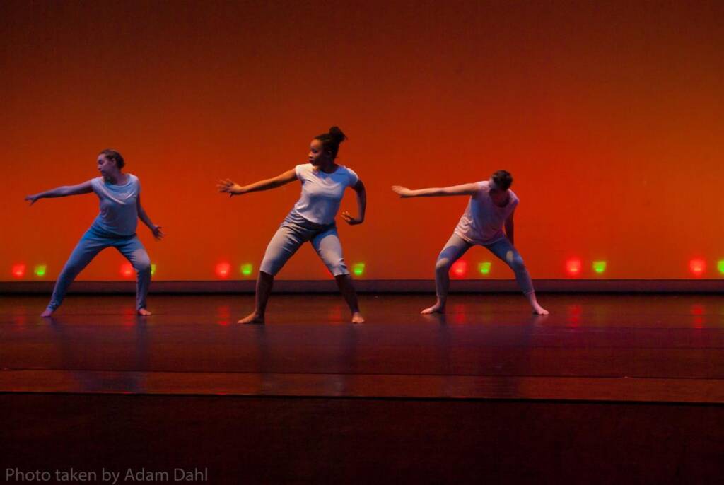 Three dancers perform on stage under warm orange lighting. They wear casual white tops and blue pants, striking dynamic poses with arms outstretched. The background is softly lit with red and green lights near the floor.