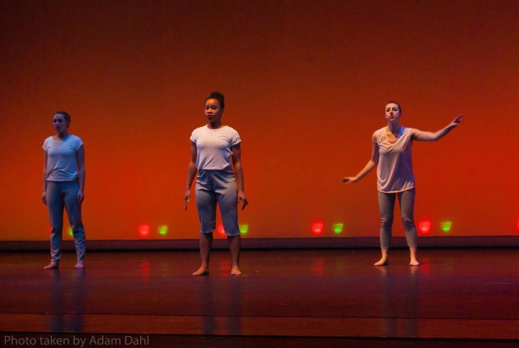 Three dancers perform on stage against a warm orange backdrop. They are wearing white tops and light-colored pants. The scene is lit with soft, colored lights along the floor. The atmosphere appears calm and focused.