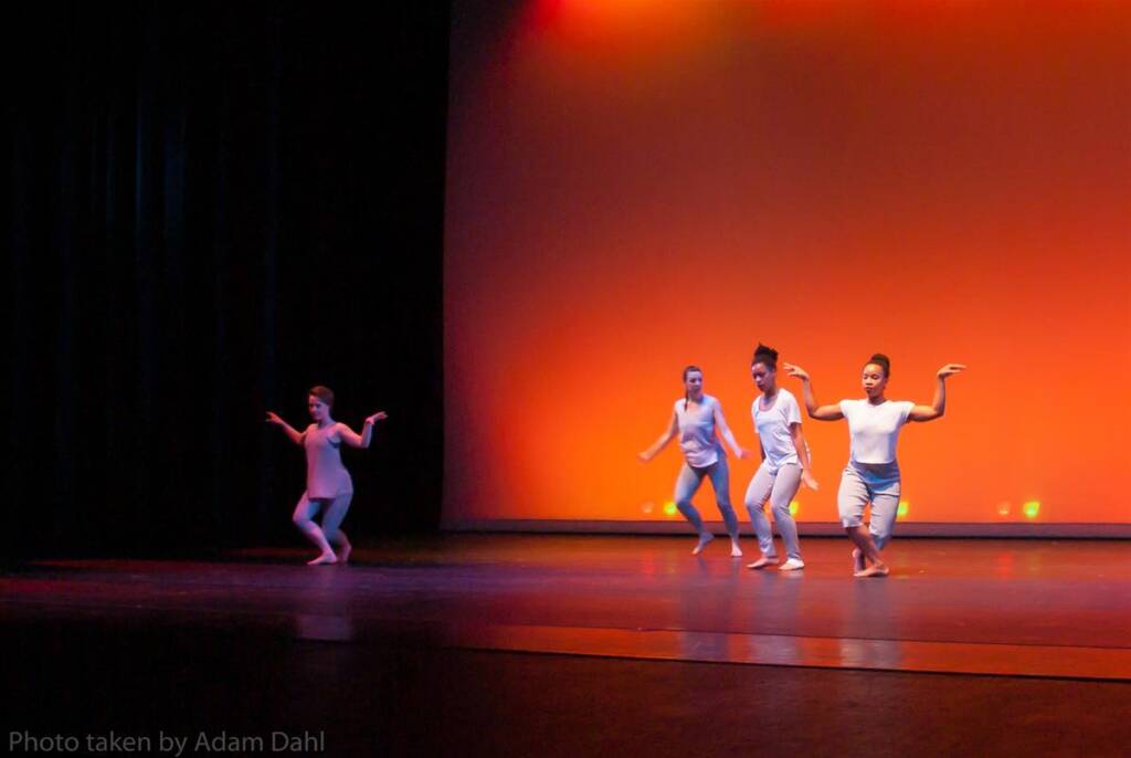 Four dancers perform on stage under warm orange lighting. They are dressed in white outfits and are in various expressive poses. The background is a smooth, illuminated wall with the stage floor dimly lit.