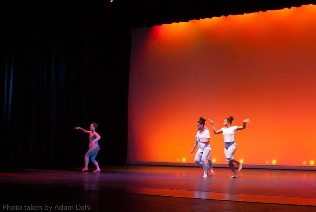 Three dancers on stage, performing against a vibrant orange backdrop. One dancer is on the left in a singular pose, while the other two on the right are in mid-motion. The stage is dimly lit with a focus on the dancers.