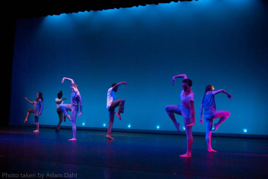 Six dancers perform on stage under blue lighting, each lifting one leg and stretching their arms in a graceful pose. They wear simple, casual dance attire, and the stage is bathed in a calming blue ambiance.