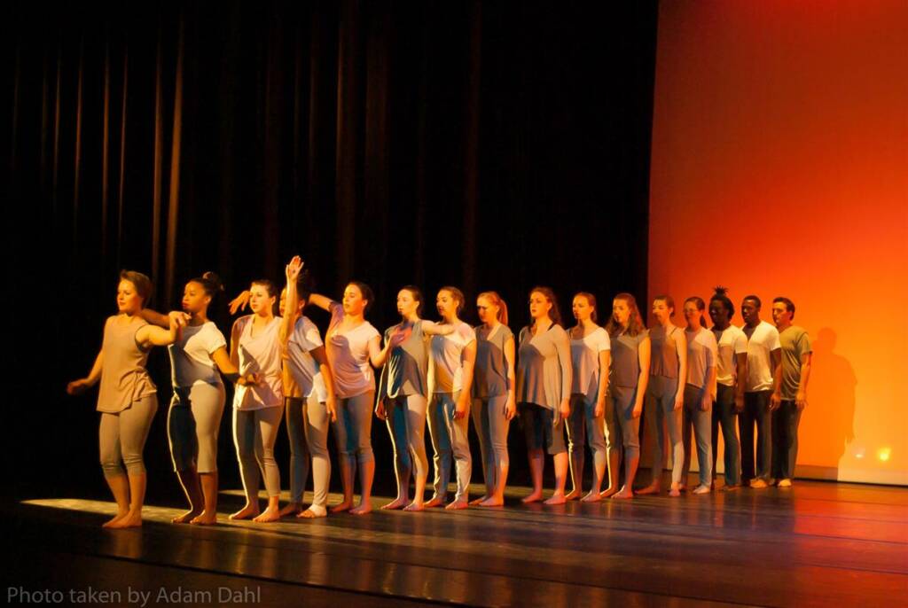 A line of dancers on stage, dressed in light gray outfits, perform synchronized movements. The background features warm orange lighting. They stand on a dark, polished floor, creating a contrast with the lit backdrop.