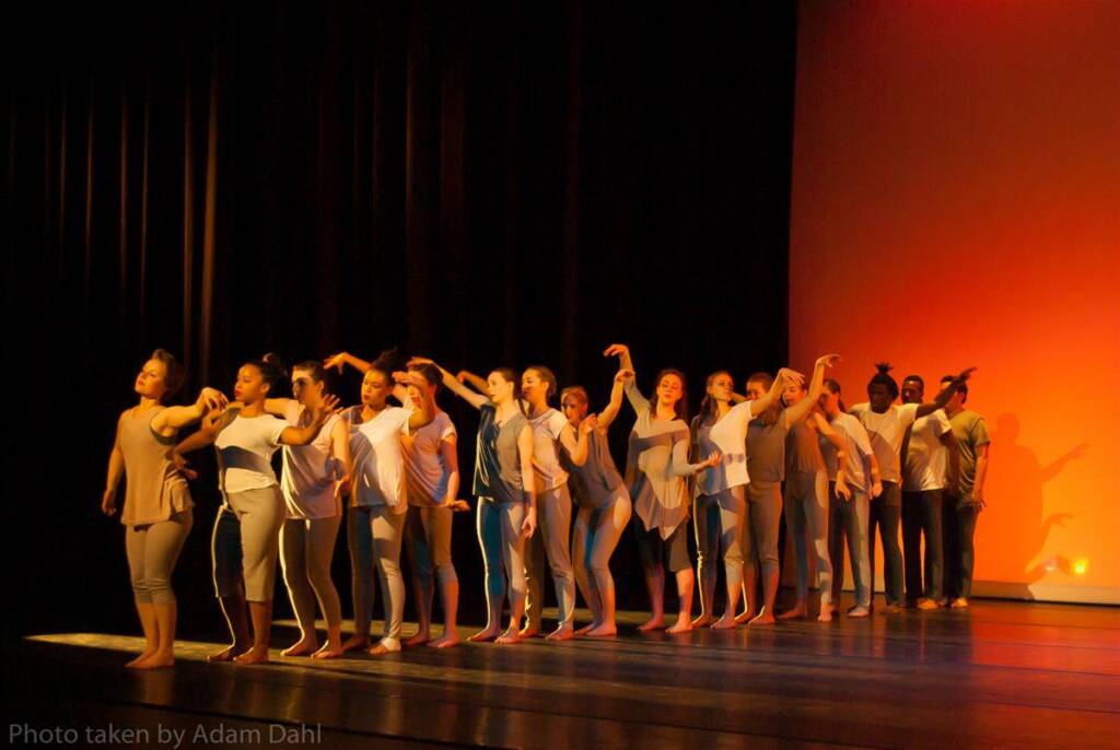 A diverse group of dancers in casual attire perform on stage under warm lighting. They stand in a line, executing a synchronized pose with arms raised and bent. The background features a gradient of dark to orange hues.