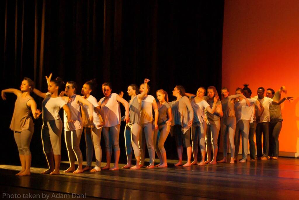 A group of dancers in matching outfits performs on stage, lined up in a single row. The backdrop is a warm orange, and the lighting casts dramatic shadows. Each dancer strikes a unique pose, showcasing a variety of expressive movements.