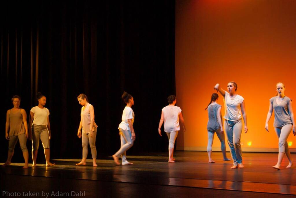 A group of dancers in casual outfits perform on stage under warm lighting. Some face forward, while others are turned away, creating a dynamic scene. The stage background is softly illuminated with an orange glow.