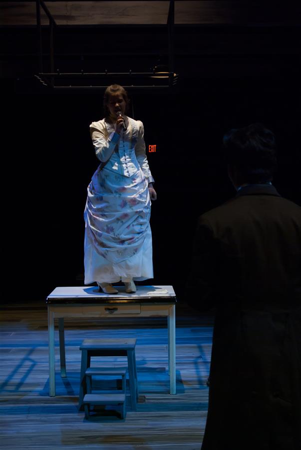 A person in a white dress stands on a table, holding a microphone, under dim stage lighting. Another person, partially visible, stands in the foreground. There's a small stool in front of the table. The scene has a dramatic atmosphere.