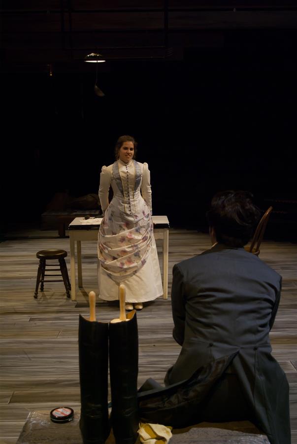 A woman in a period dress stands near a table, facing a seated person in a suit on a stage set. The scene is dimly lit with wooden flooring and theater props, including a pair of boots in the foreground.