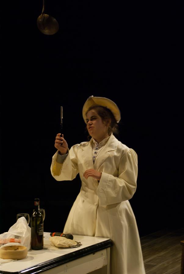 Person in a white coat and hat examines a kitchen tool, possibly a peeler, with a thoughtful expression. They stand next to a table with various kitchen items, including a wine bottle and a loaf of bread, against a dark background.