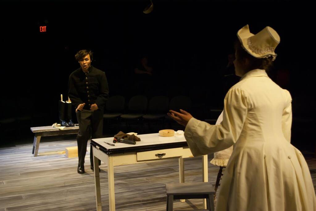 Two actors perform on stage. One wears a dark uniform, standing by a table with props like a brush and bowl. The other wears a white bonnet and dress, facing away. Wooden chairs and dim lighting suggest a dramatic scene.