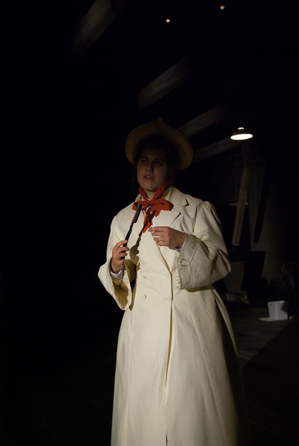 A person stands in a dimly lit area, wearing a long cream coat and a wide-brimmed hat. They hold a string tie in their hands. The background is mostly dark with some visible lights and structures.