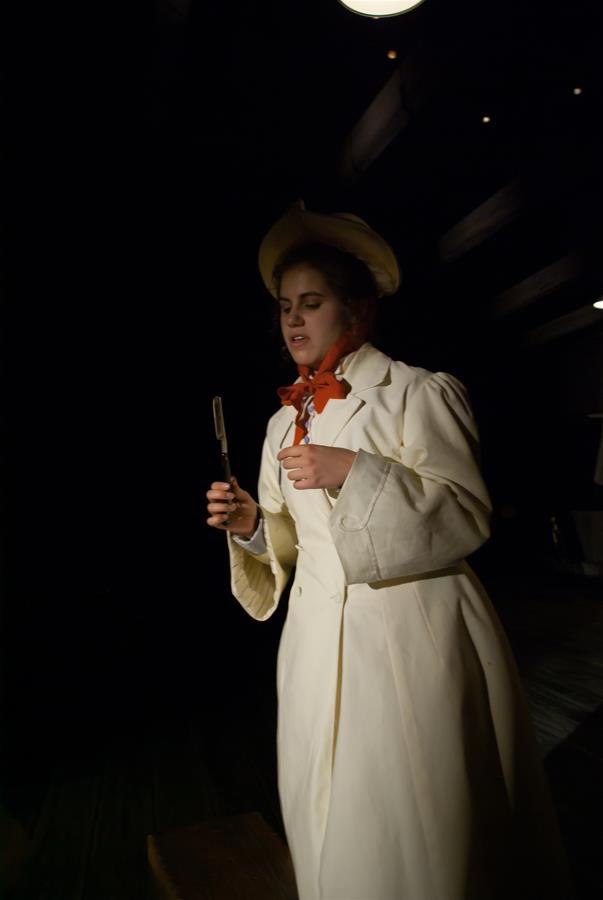 Person in a light-colored coat and hat holding a folding knife with a red scarf, standing against a dark background.