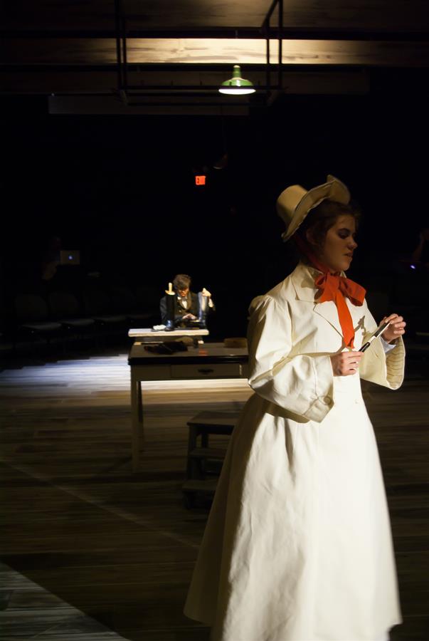 A person in a vintage white outfit and hat, holding glasses, stands on a dimly lit stage. A second person is seated at a wooden desk in the background under a spotlight, working with papers. The scene has a theatrical setting.