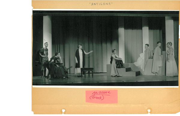 A black and white photo of a theatrical performance of "Antigone." Actors in ancient Greek costumes are on stage. One actor stands to the left, gesturing, while others are seated or standing with props on a simple set with columns.